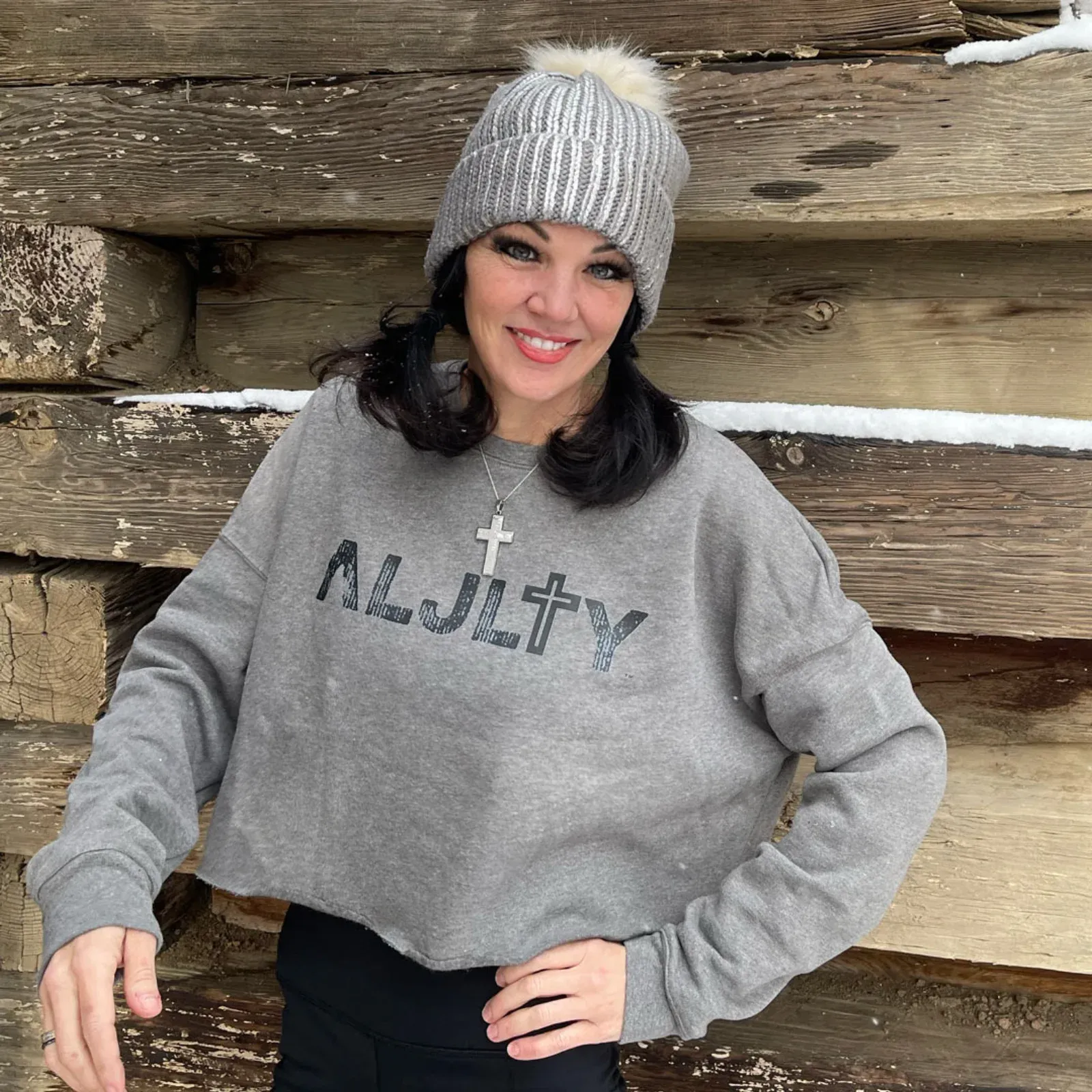 Elizabeth wearing a gray ALJLTY cropped sweater and knit beanie, standing against a rustic log wall with snow in the background